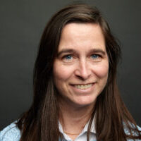 Professional portrait of a smiling woman with long brown hair against a dark background.